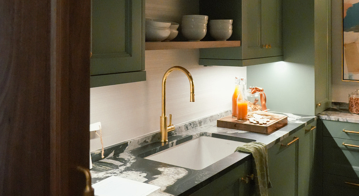 Modern kitchen with green cabinets and under-cabinet lighting, a stone countertop with a gold faucet, and juice in a glass pitcher.