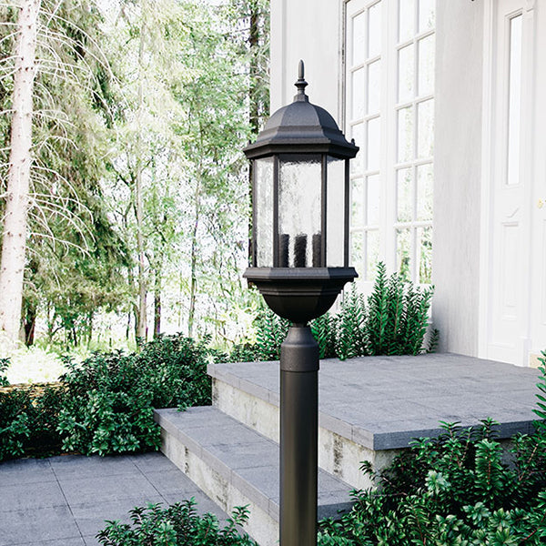Decorative outdoor lamp post on a patio with greenery and a building in the background