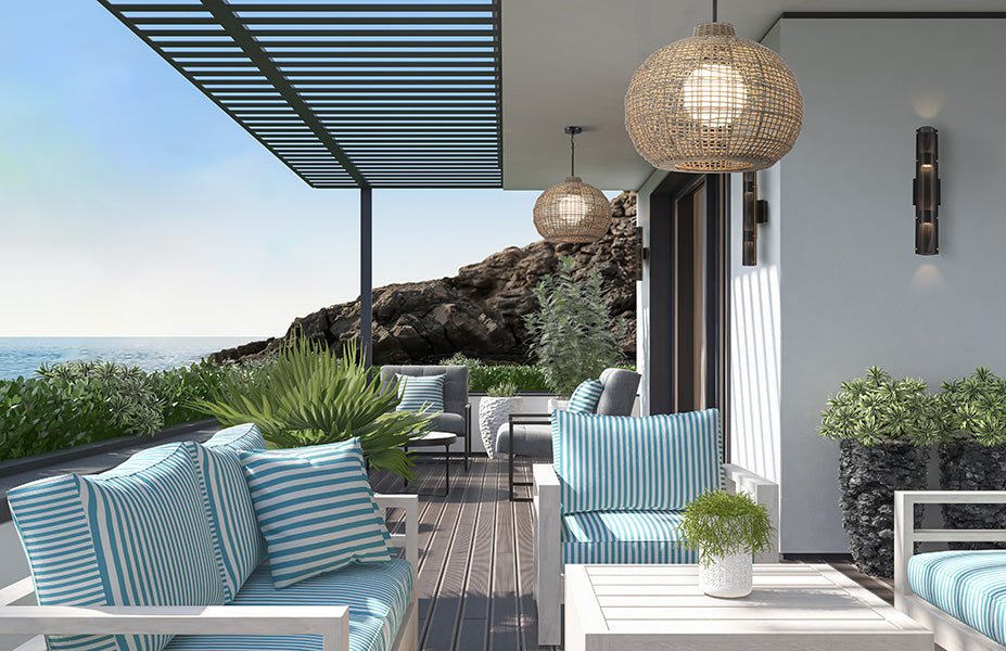 Modern outdoor patio seating area with white-framed sofas and chairs, blue-and-white striped cushions, woven pendant lights, and a coastal rock-and-sea backdrop.