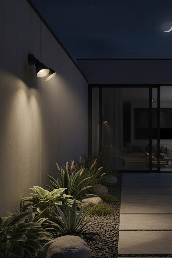 Side of a house with a wall-mounted light fixture illuminating plants and a walkway at night.