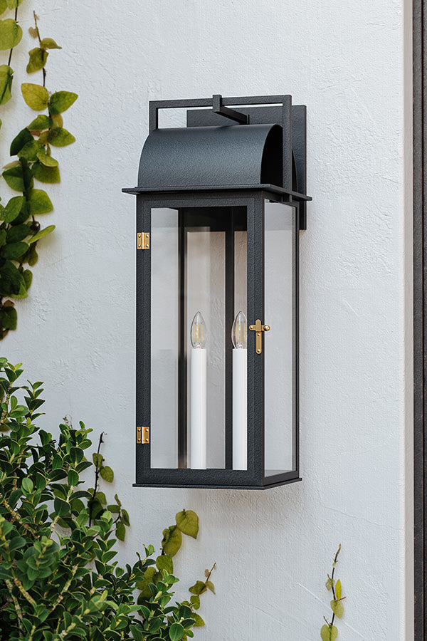 Black outdoor wall lantern with two candles on a white wall, surrounded by greenery.