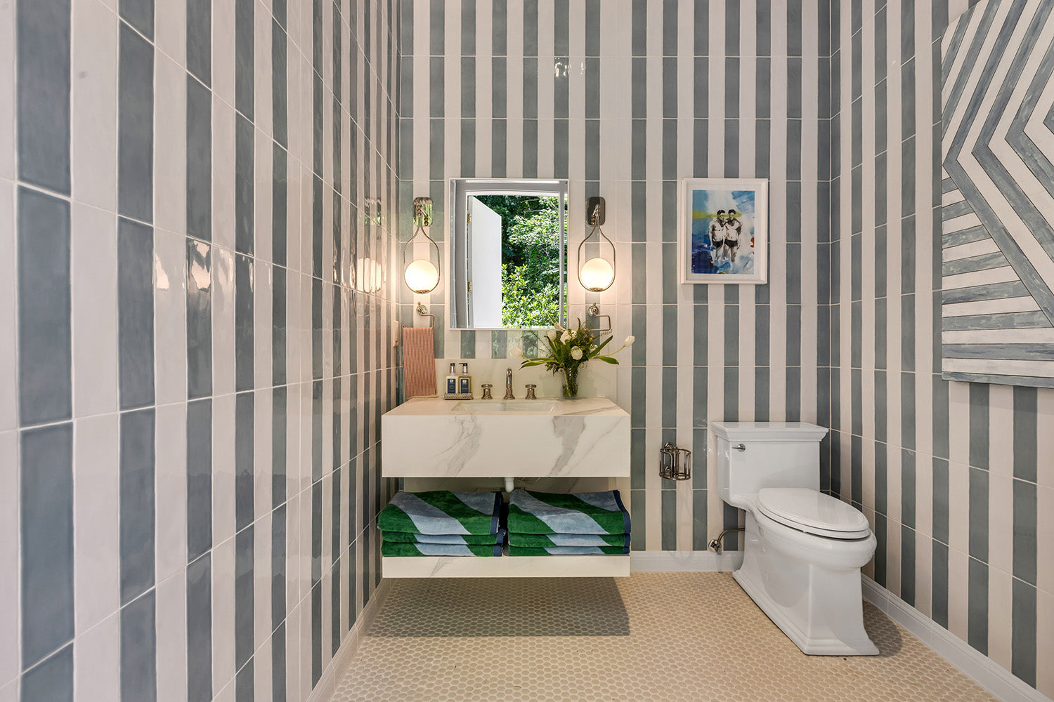 Pool house bathroom with striped tile walls, a floating vanity, two wall sconces, and a Kohler toilet.