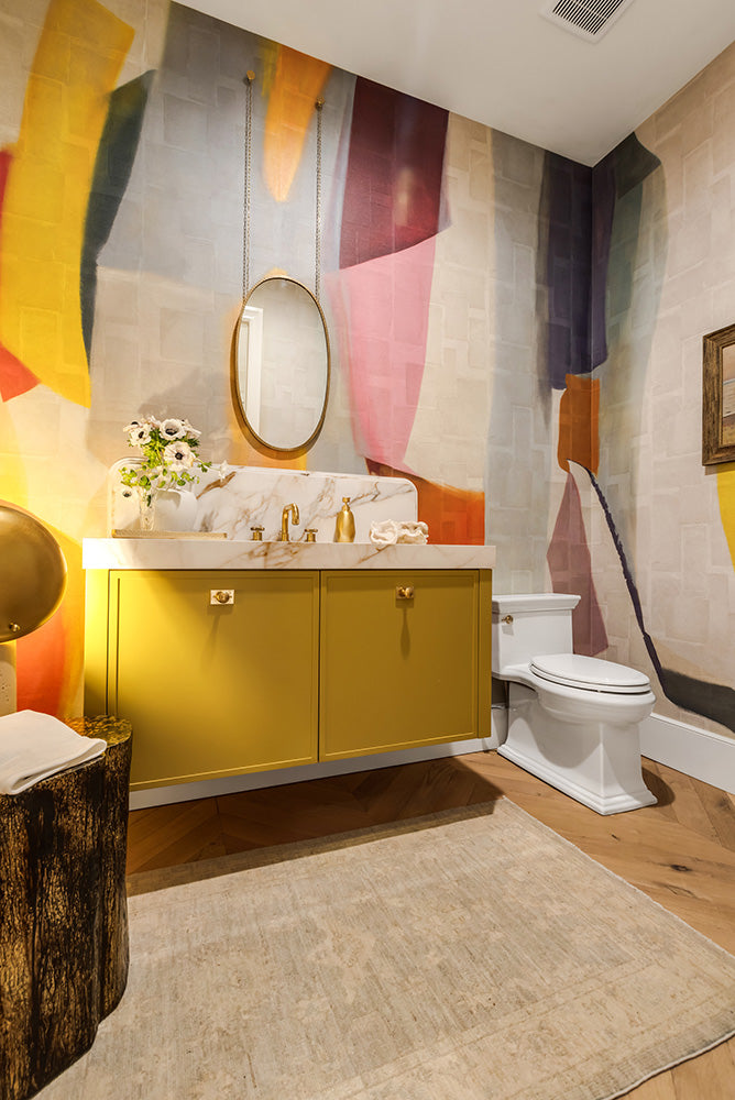 Bold, colorful guest bathroom with abstract wallpaper, a deep yellow floating vanity with gold faucets and a hanging oval mirror. 